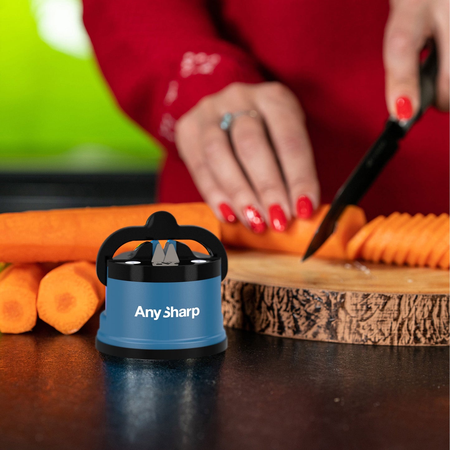 Knife sharpener labeled "AnySharp" on a kitchen counter while a person with red nails slices carrots on a wooden board in the background.