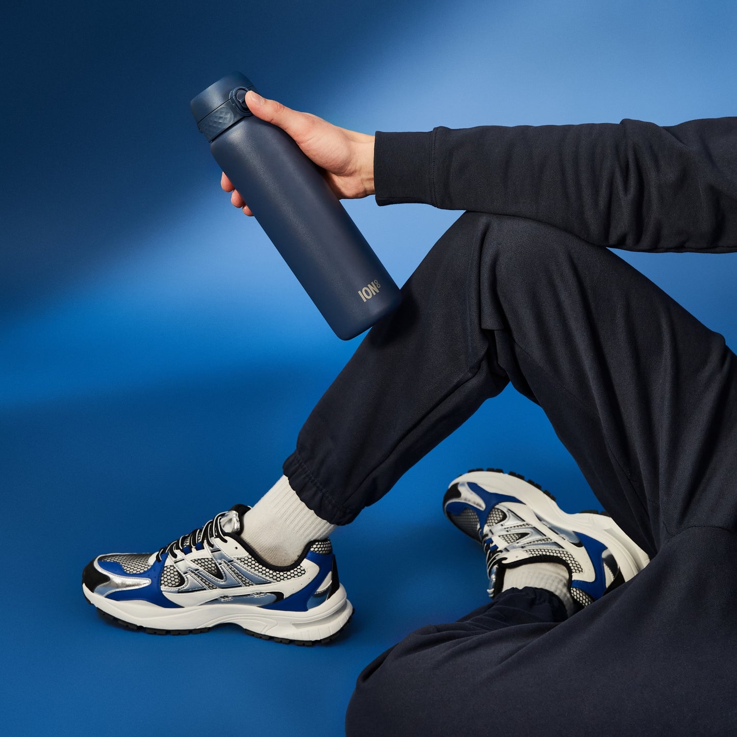 A person holds a dark-blue ION8 water bottle. They are seated, wearing dark sweatpants and sneakers, against a blue backdrop.