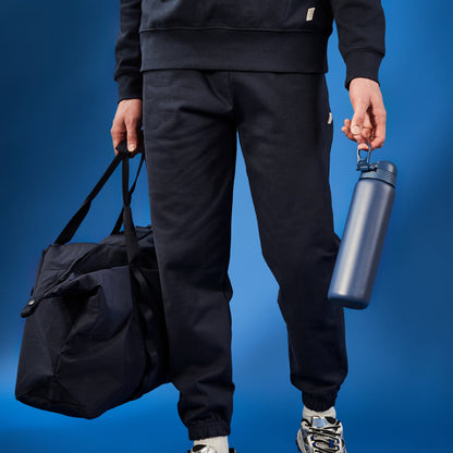 A person carries a dark duffel bag and a dark blue water bottle against a blue backdrop.