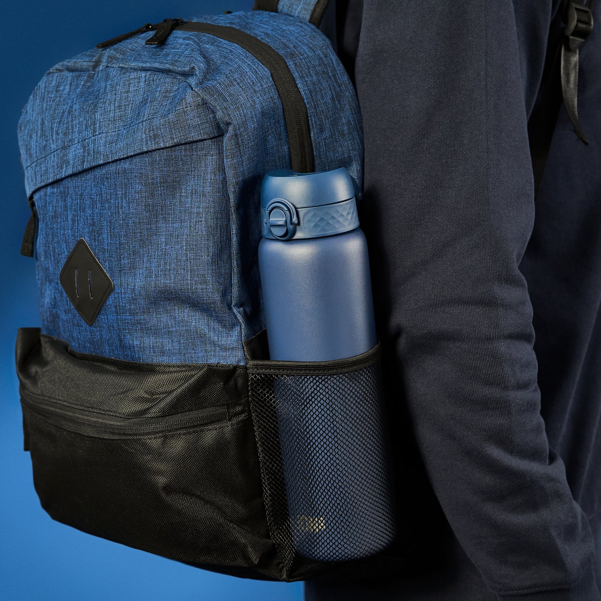 A blue water bottle is held in a backpack's side mesh pocket. The backpack is worn by someone in a dark blue sweatshirt against a blue background.