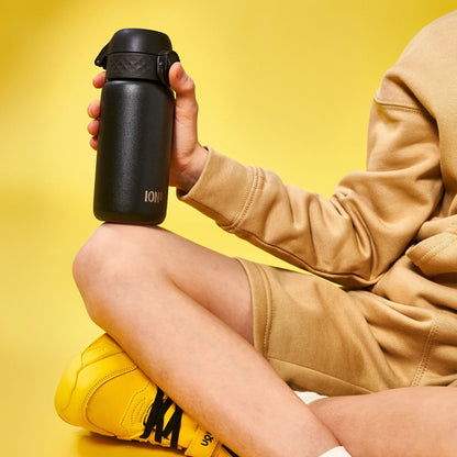 A person holds a black water bottle labeled ION8 while sitting cross-legged wearing yellow sneakers in a bright yellow environment