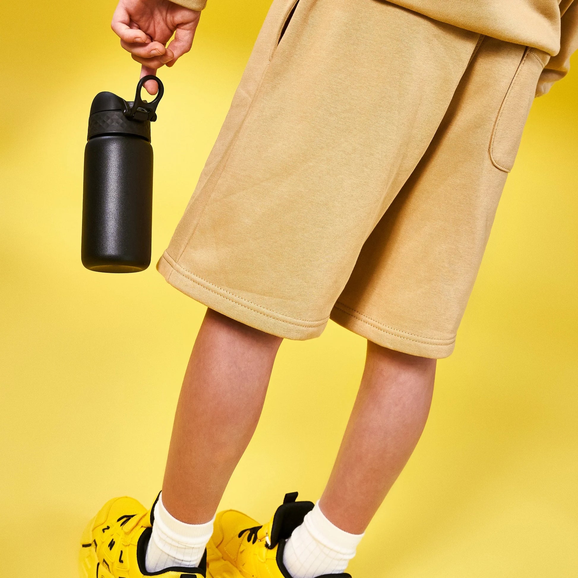 A person holds a black water bottle against a yellow background wearing tan shorts and yellow sneakers with white socks.