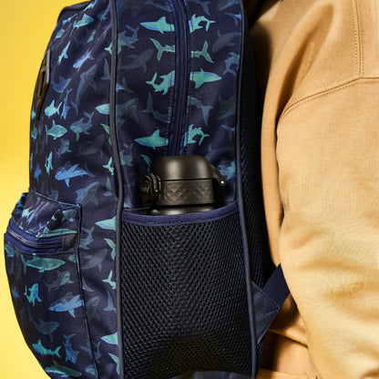 A navy backpack with blue shark pattern holds a black water bottle in its side mesh pocket worn by a person in a tan hoodie against a yellow background.