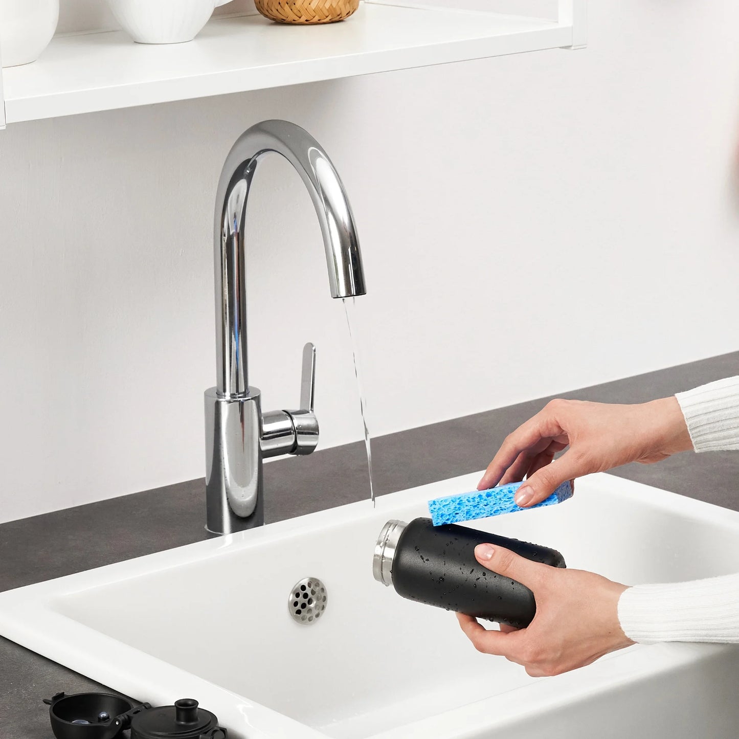 Hands scrub a black water bottle with a blue sponge under running tap water in a modern kitchen sink environment.