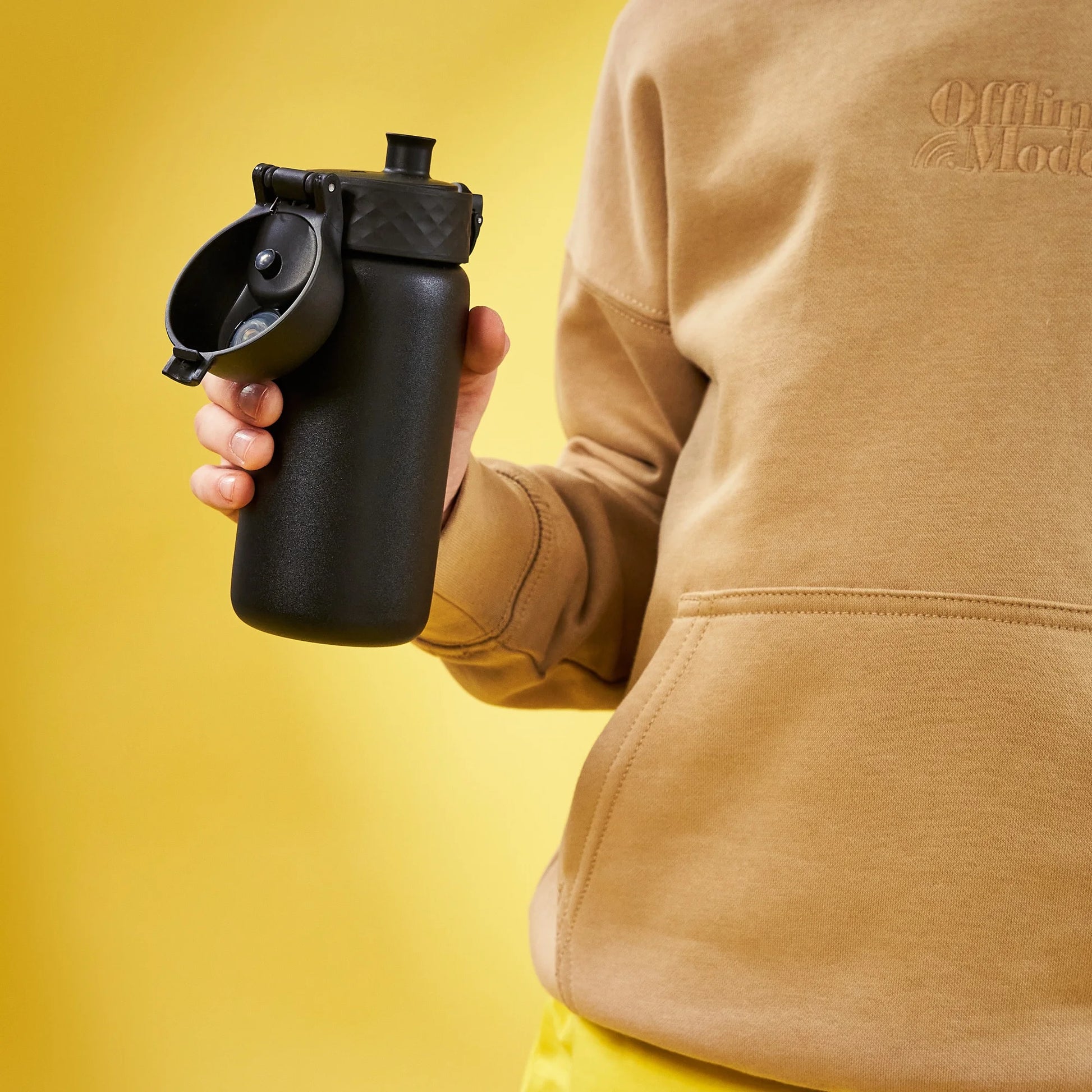 Hand holding a black insulated water bottle with a flip-top lid wearing a tan sweatshirt in front of a solid yellow background showing the text "Offline Mode" on the sweatshirt