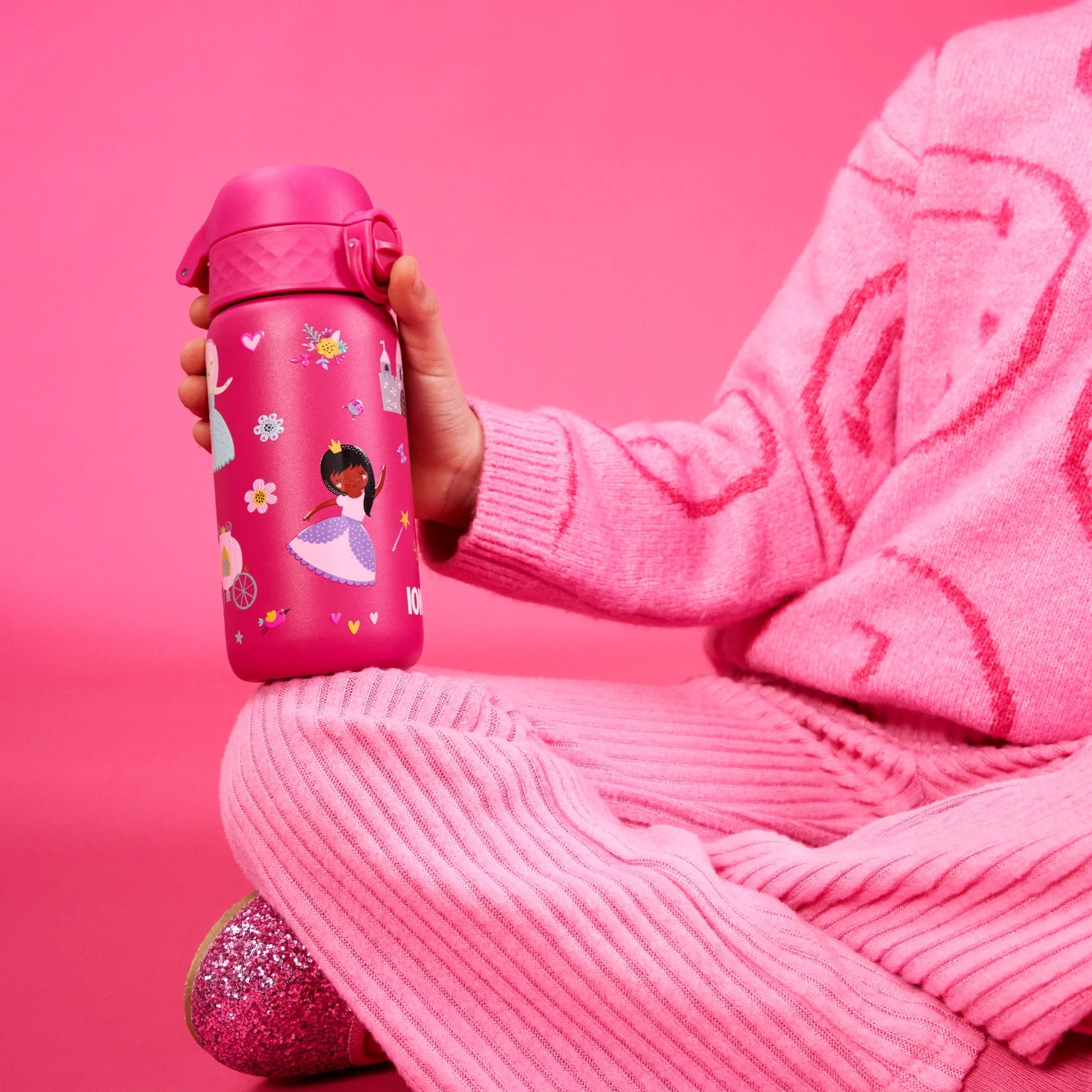 A child holds a pink water bottle decorated with princess and floral illustrations while sitting cross-legged in pink knit clothing against a pink background.