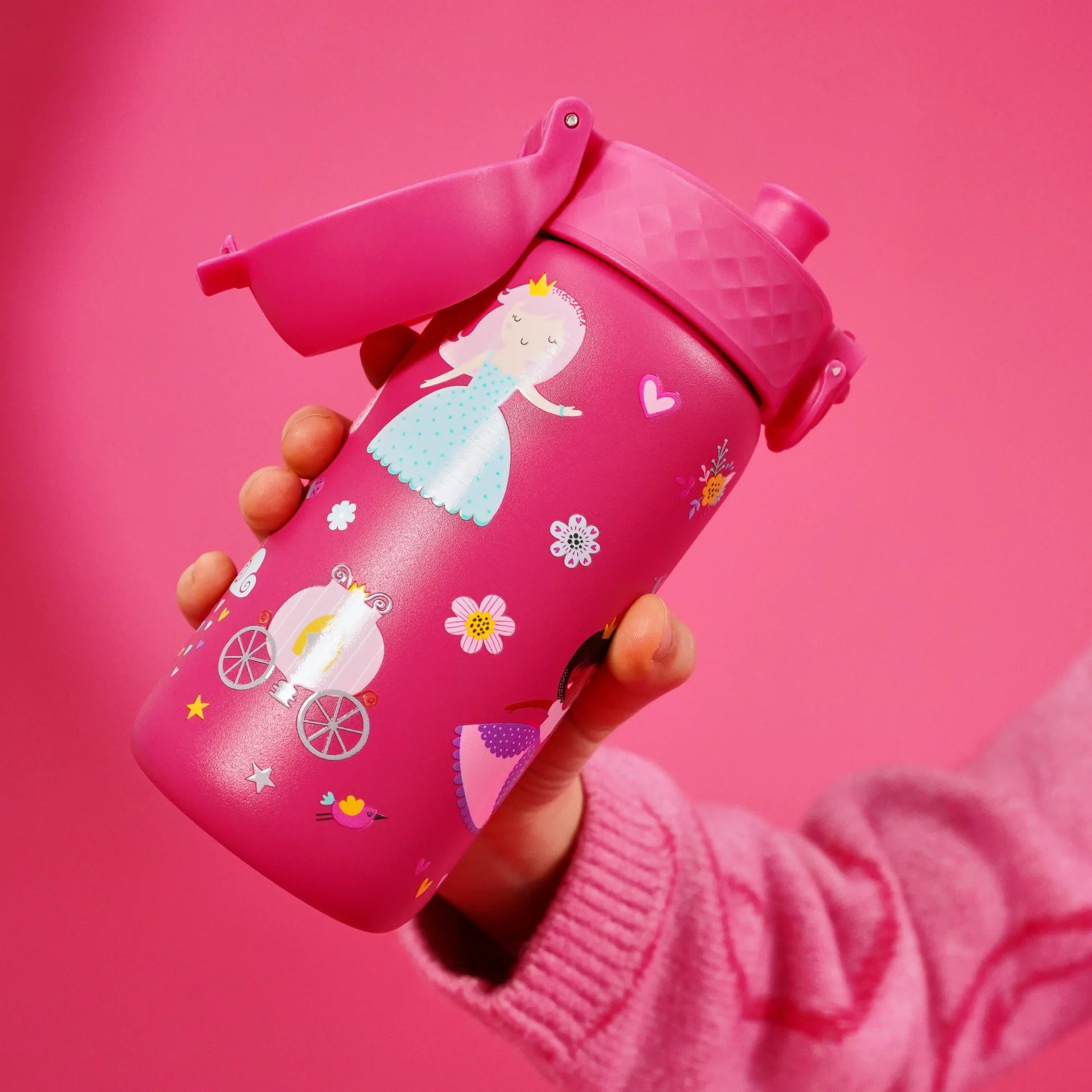 A child’s hand holds a pink water bottle decorated with a fairy tale theme featuring princesses, a carriage, flowers, and hearts, set against a matching pink background.