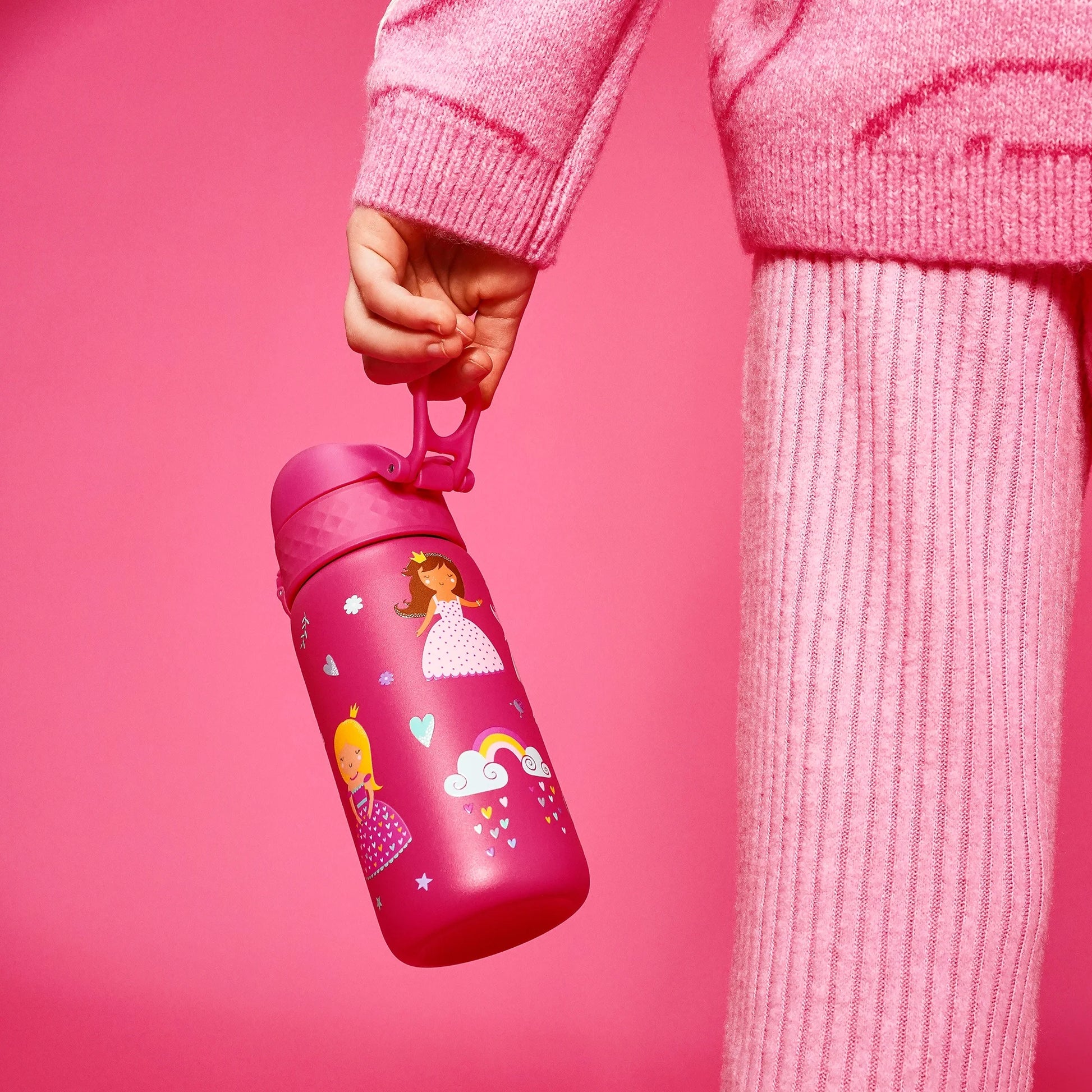 A child's hand holds a pink water bottle featuring princess and rainbow illustrations against a matching pink outfit and background.