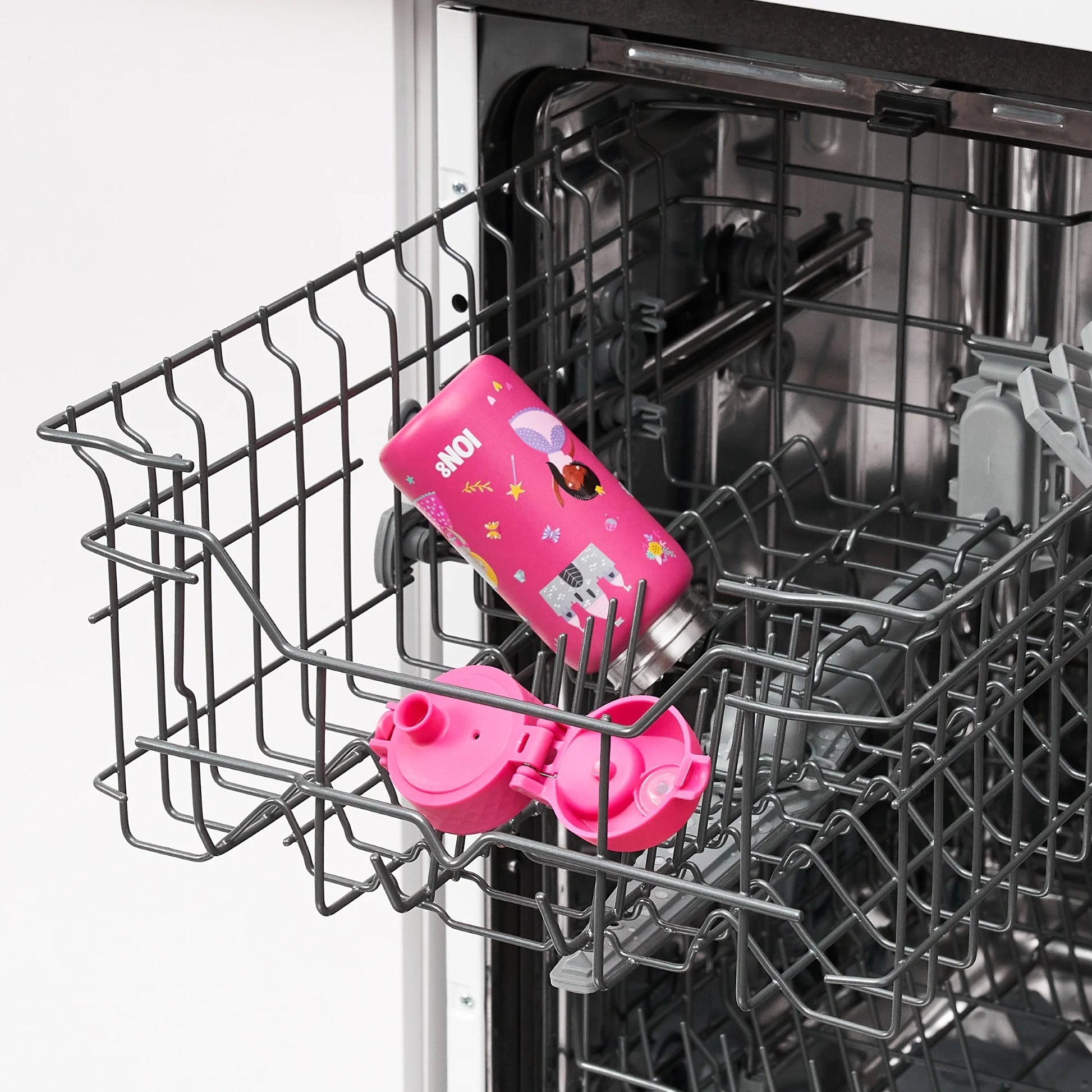 A colorful pink water bottle with a unicorn design lies on its side in an open dishwasher rack surrounded by gray metal prongs in a kitchen setting