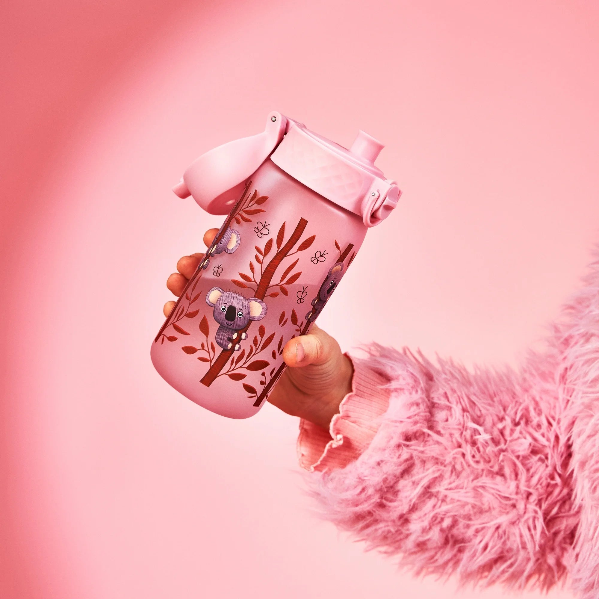 A pink water bottle with a koala design is held by a child wearing a pink fuzzy sweater against a pink background.