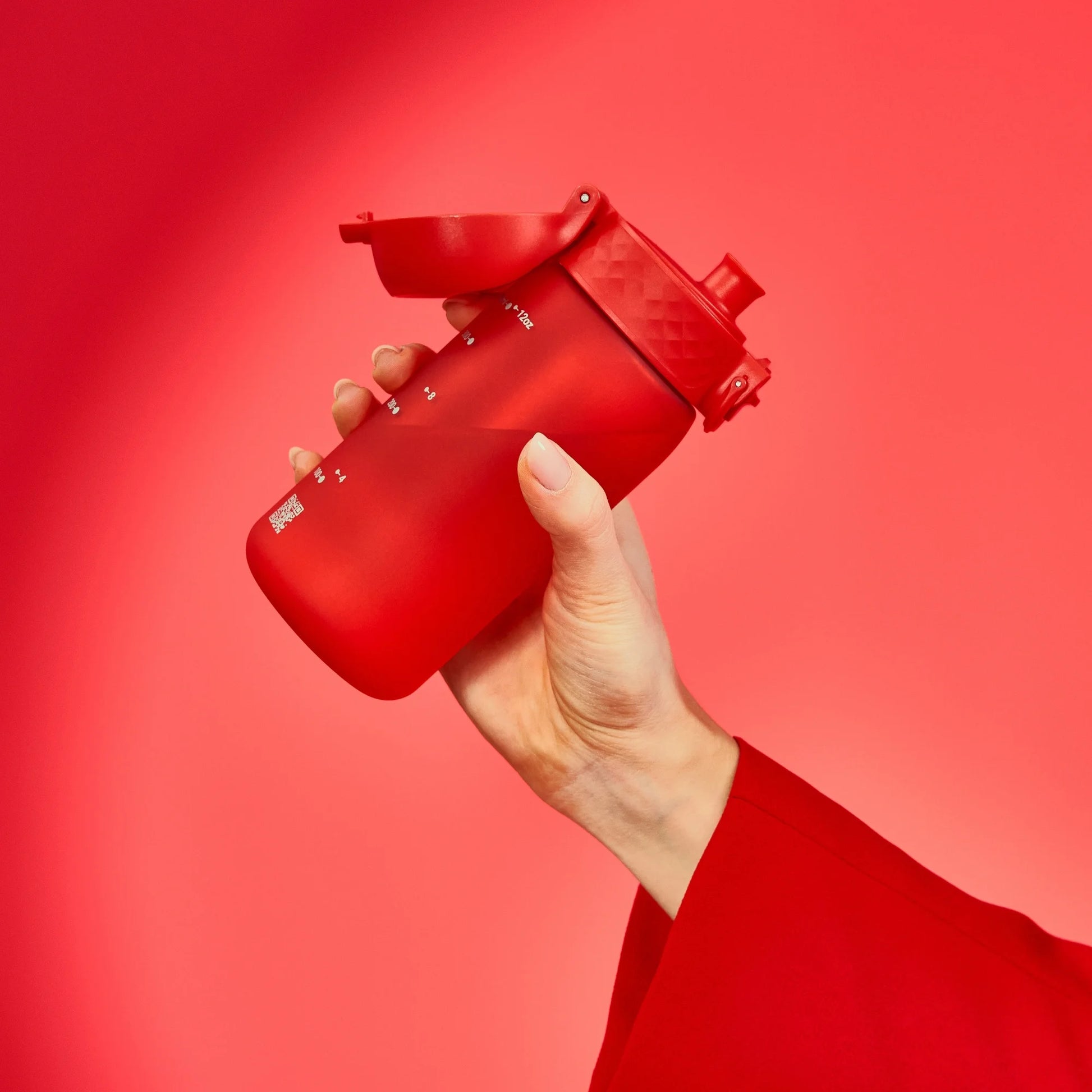 A red sports bottle with a flip cap is held by a hand against a matching red background. The bottle features measurement markings with labels like 10 oz and 20 oz.