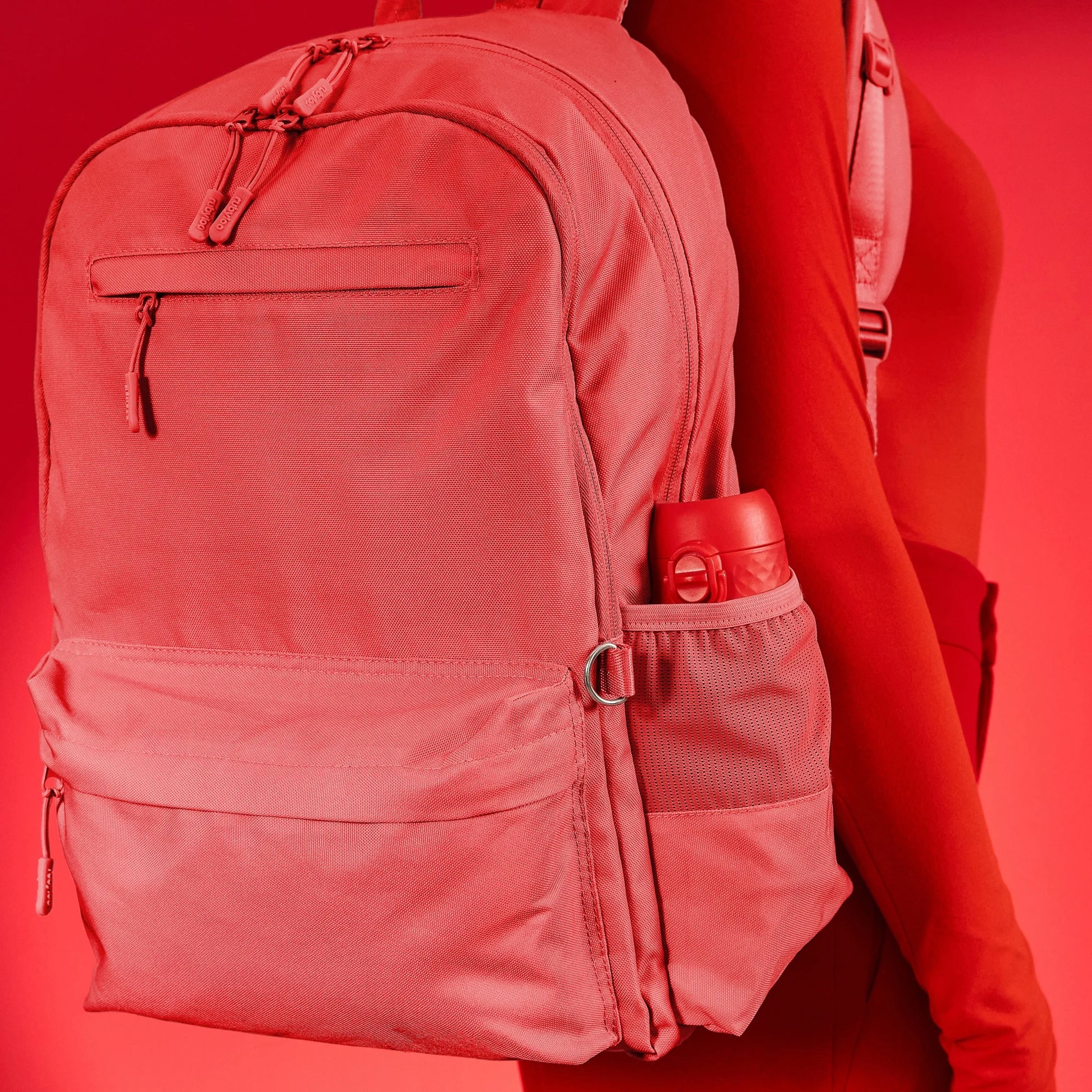 A red backpack with multiple compartments and zippers is worn on a person’s back. A red water bottle is in the side pocket against a red background.