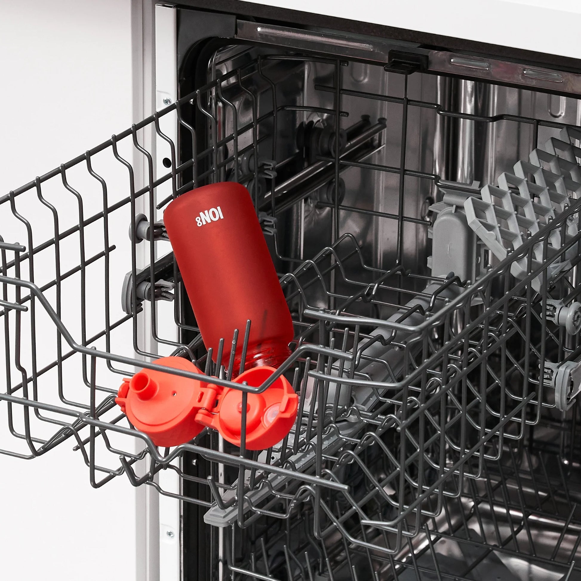 A red bottle with white text "ION8" is positioned on its side in the lower rack of an open dishwasher surrounded by metal prongs and grey plastic elements.