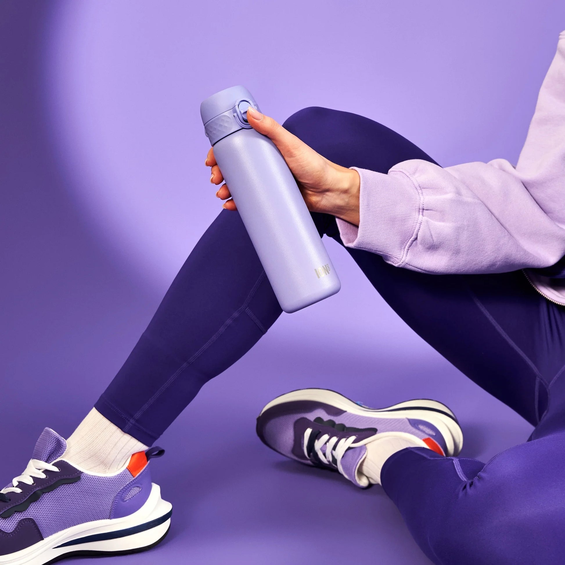 Hand holding a purple water bottle labeled "LARQ" resting on lavender leggings with purple sneakers in a purple-themed studio setting