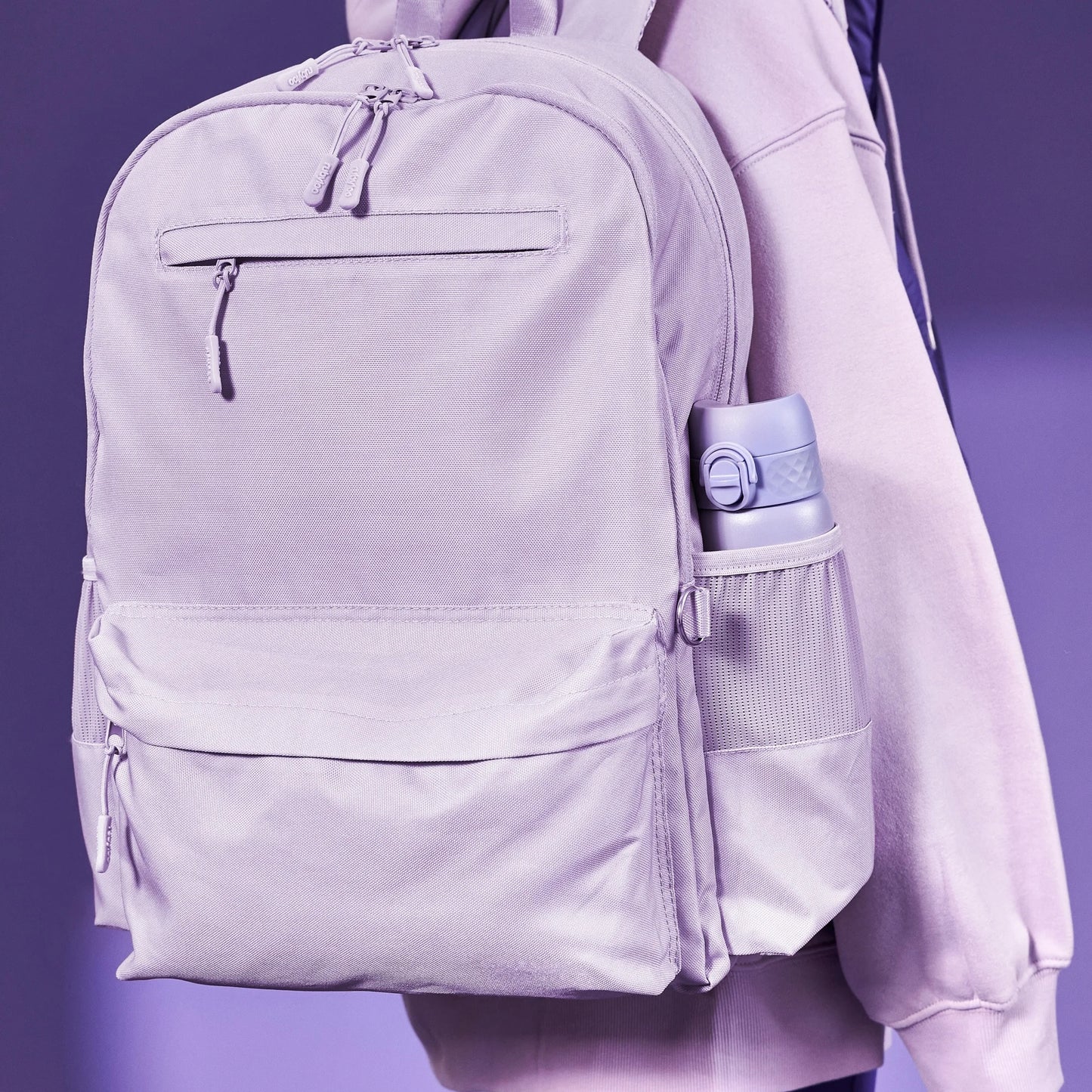 A lavender backpack with multiple zippers hangs from the shoulder of a person wearing a matching hoodie; a water bottle is in the side pocket against a purple background.