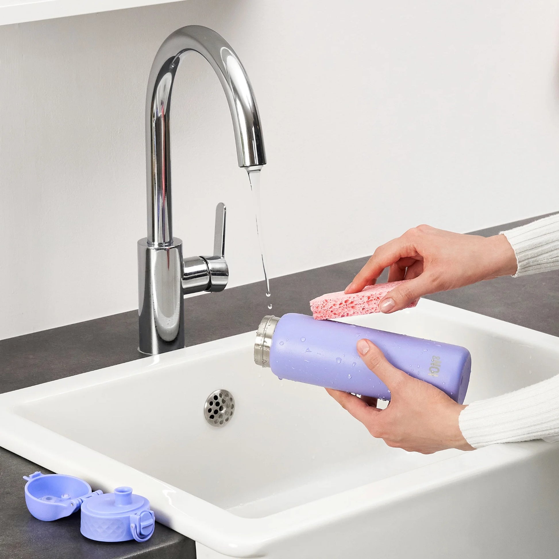 Hands clean a purple water bottle with a pink sponge under a running faucet in a kitchen sink context with a removed blue cap nearby.