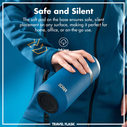 A person's hand holds a blue ION8 travel flask with a soft base for silent placement. Text reads: "Safe and Silent The soft pad on the base ensures safe, silent placement on any surface, making it perfect for home, office, or on-the-go use. Soft base TRAVEL FLASK."