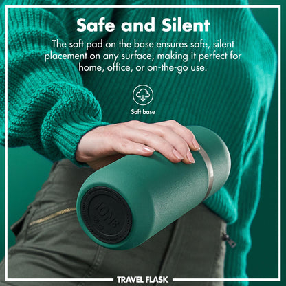 A hand holds a green travel flask with a soft base in front of a person in a green sweater and pants. Text reads Safe and Silent The soft pad on the base ensures safe silent placement on any surface making it perfect for home office or on-the-go use. Soft base TRAVEL FLASK.