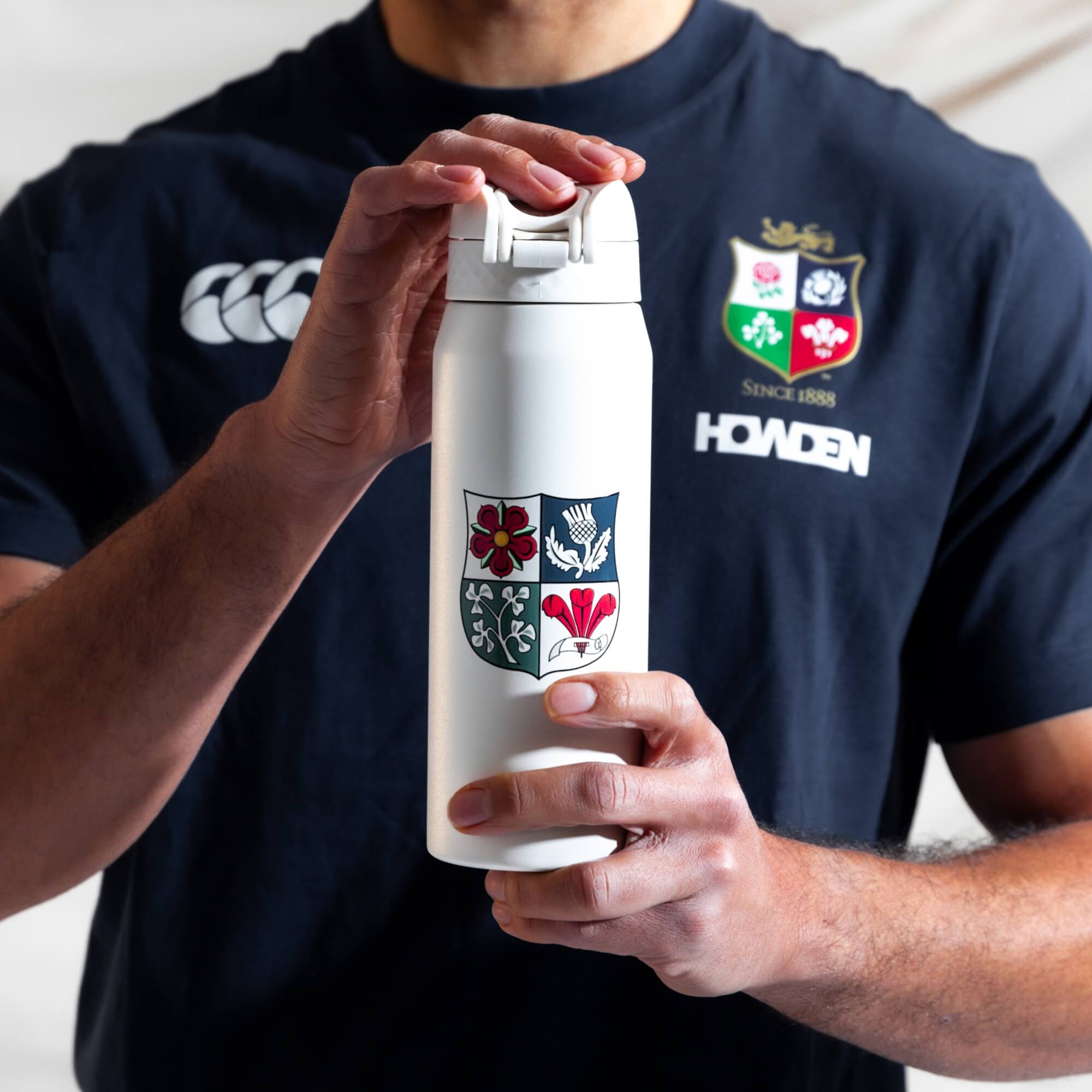 A person holds a white water bottle featuring a crest; the bottle is being opened. The person is wearing a dark rugby shirt with a logo that includes "SINCE 1888" and "HOWDEN".