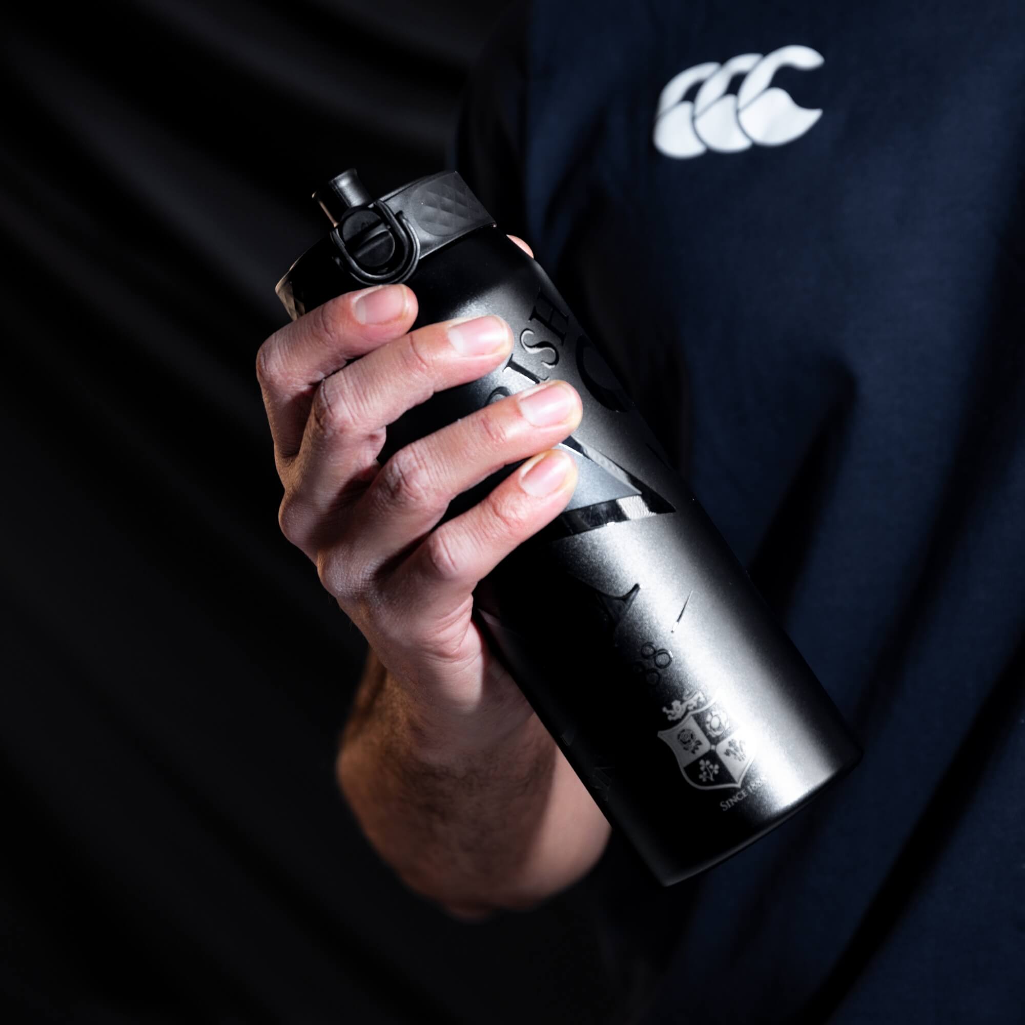 A person holds a dark gray, cylindrical water bottle with a black cap and etched design, including a crest and the text "SINCE 1888," against a dark background.