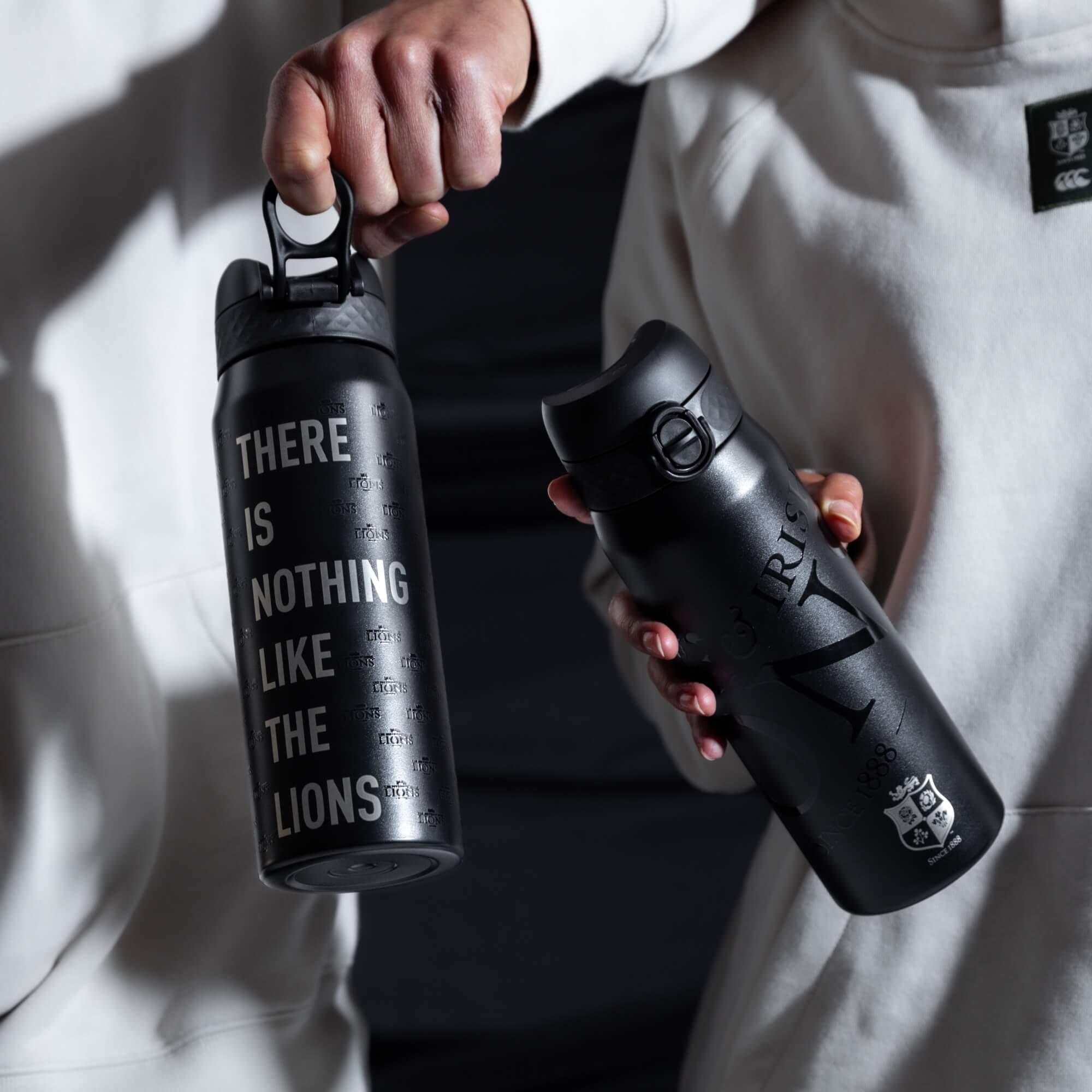 Two black water bottles are being held. One says “THERE IS NOTHING LIKE THE LIONS,” the other has “CHRIS” and “SINCE 1888” with a crest. The setting is dark, with the bottles held against light-colored clothing.