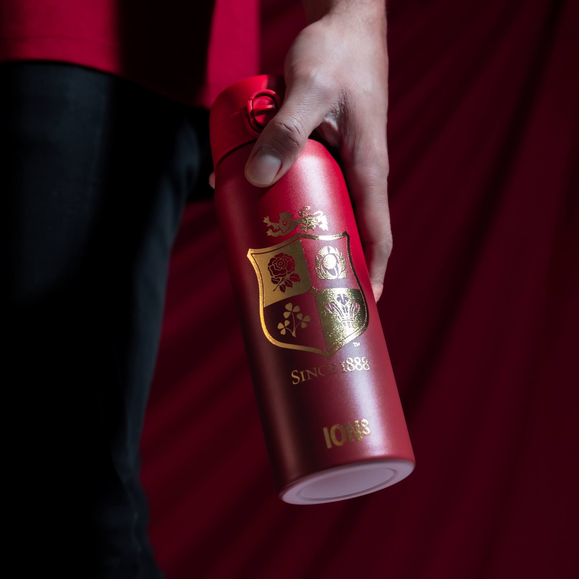 A red thermos bottle, being held, against a red background. The bottle features a gold emblem and the text "SINCE 1888" and "IONS".