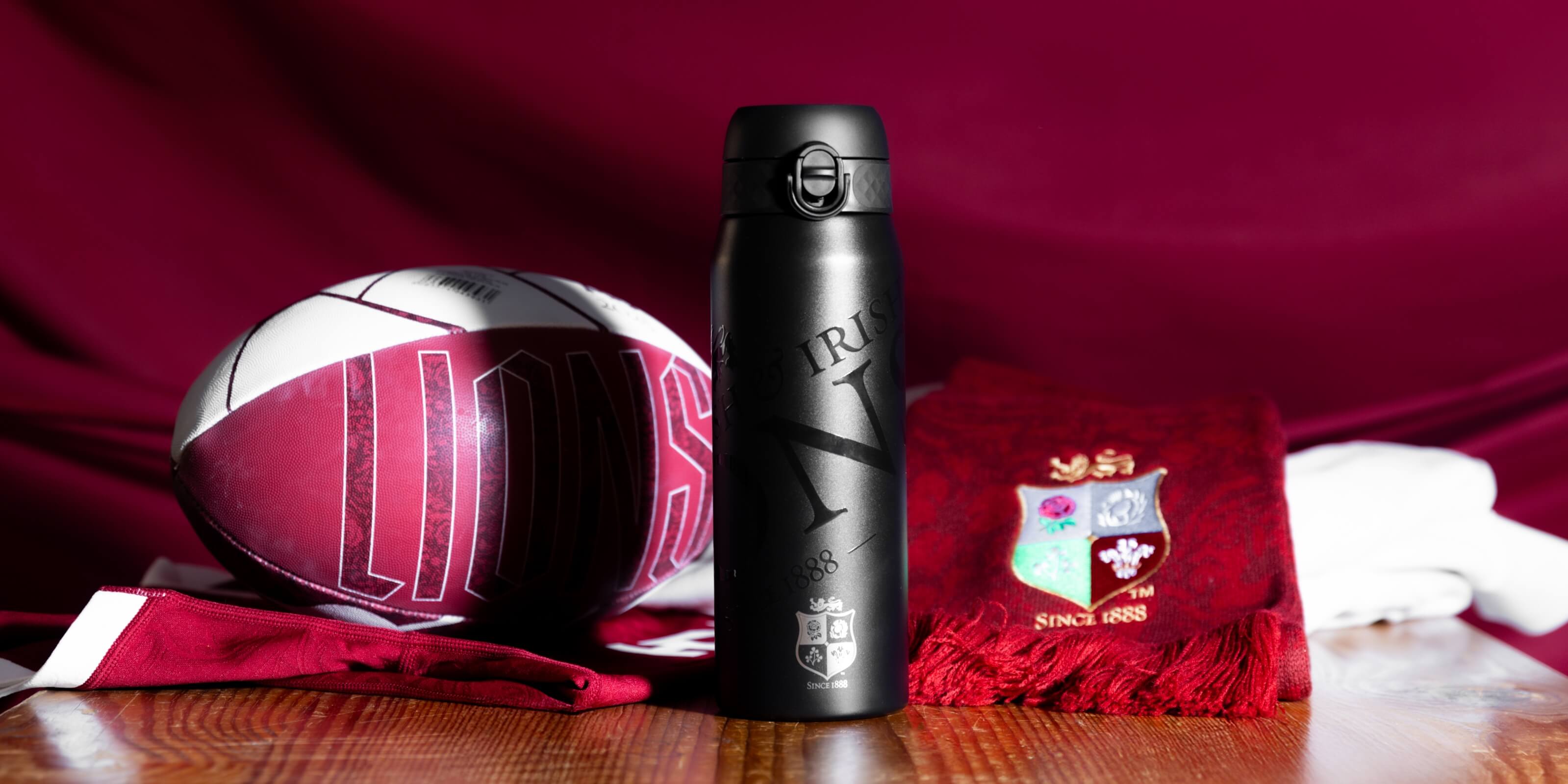 A black insulated water bottle stands near a rugby ball and scarf on a wooden surface. The bottle is engraved with "IRISH" and "1888," along with a crest and "SINCE 1888." The ball and scarf also feature team branding.