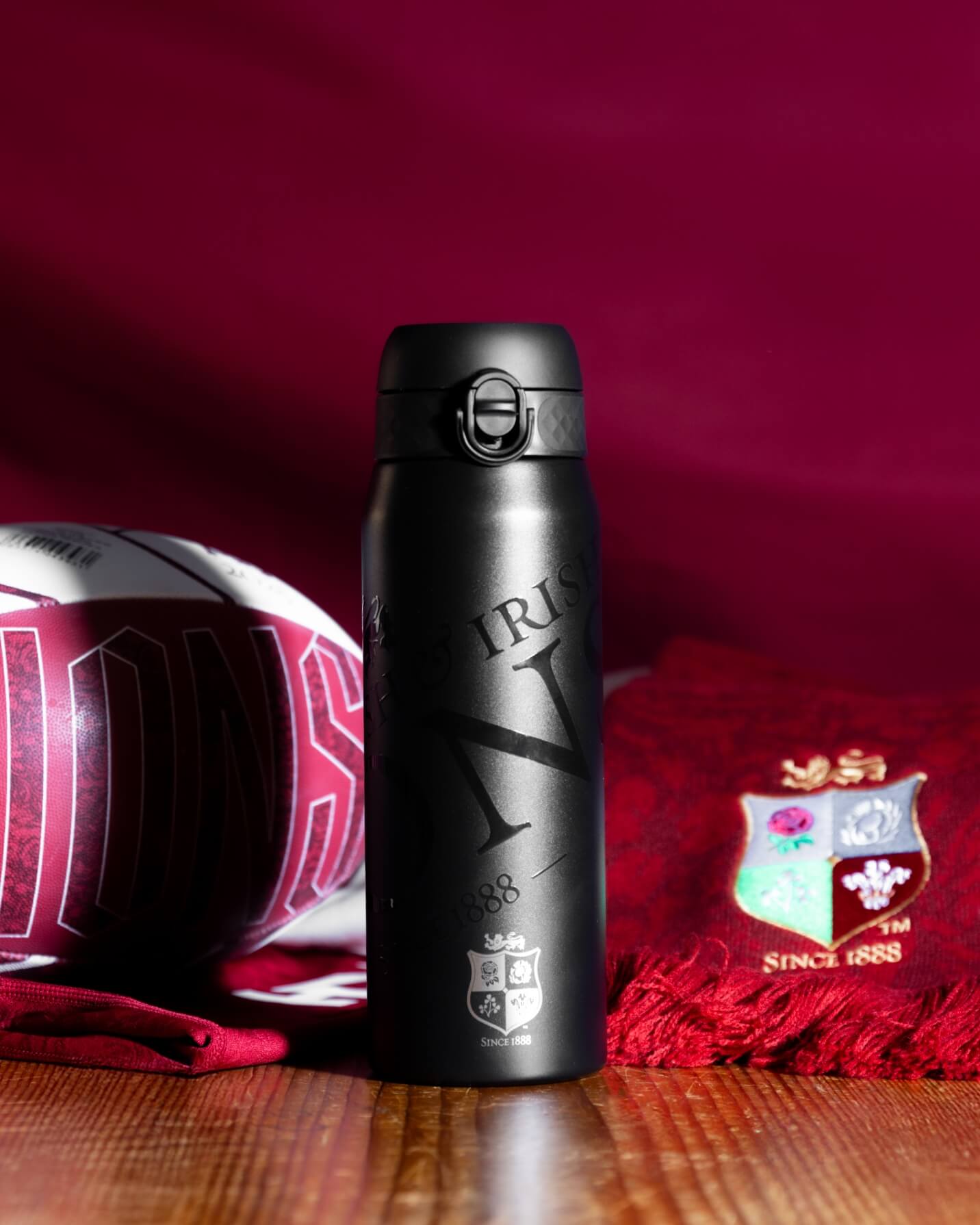 A black water bottle stands on a wooden surface. It features engraved text: "IRIS," "N," "1888," and a crest with "SINCE 1888." A rugby ball and a burgundy scarf with a similar crest are nearby.