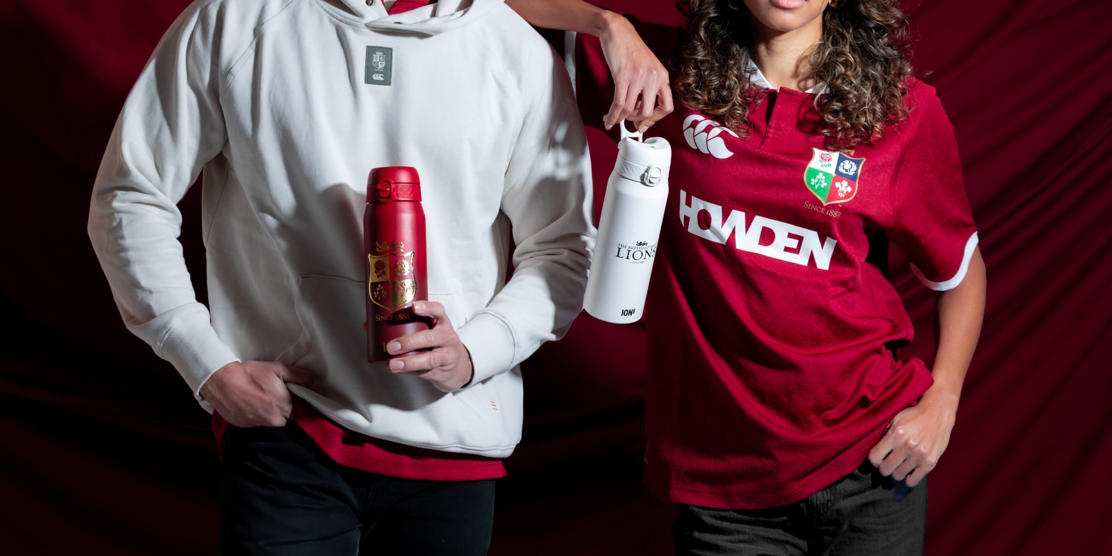 Two individuals hold insulated water bottles against a dark red backdrop. One bottle is red with gold detailing, the other is white with black text. The text on the white bottle reads: “THE BRITISH & IRISH LIONS” and “ION®”. The red jersey has “HOWDEN” and “SINCE 1888” printed on it.