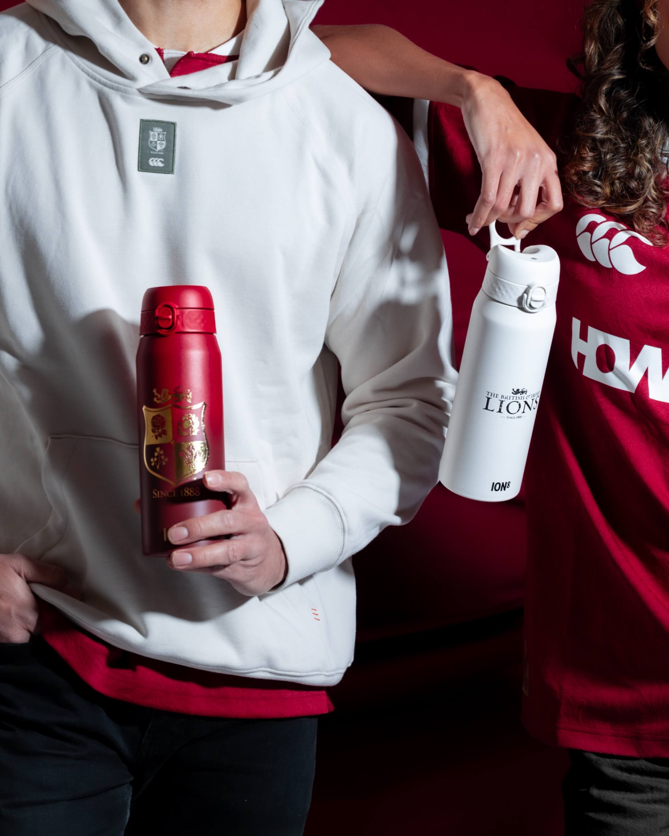 Two people hold insulated water bottles. One bottle is red, the other white. Both bottles display the British & Irish Lions logo; the white one also displays “ION8” and “SINCE 1888”. The setting is dark, with a burgundy backdrop.