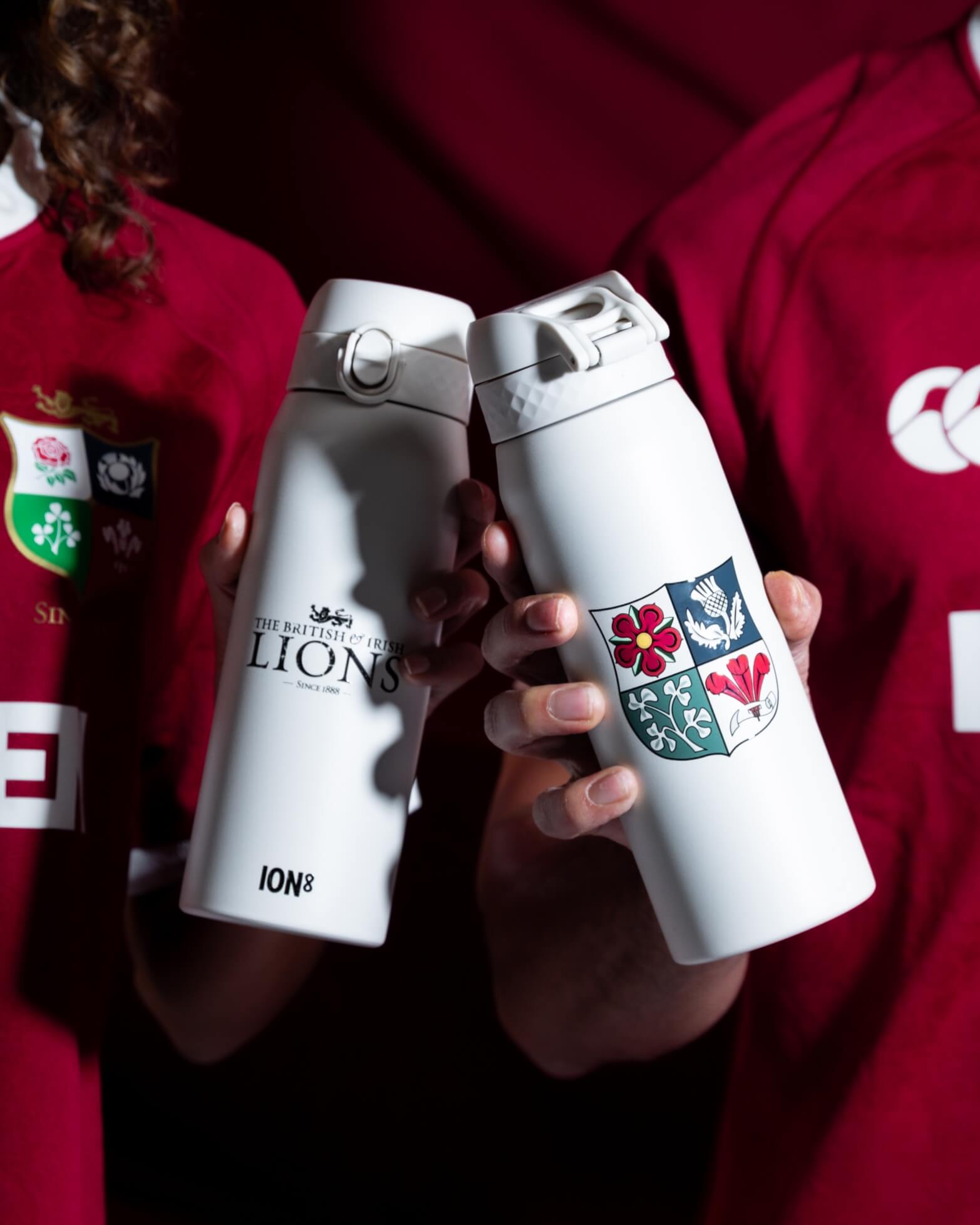 Two white water bottles, one displaying "THE BRITISH & IRISH LIONS - SINCE 1888," the other with a heraldic emblem, are held by individuals wearing red rugby jerseys. ION8 is printed on one bottle.