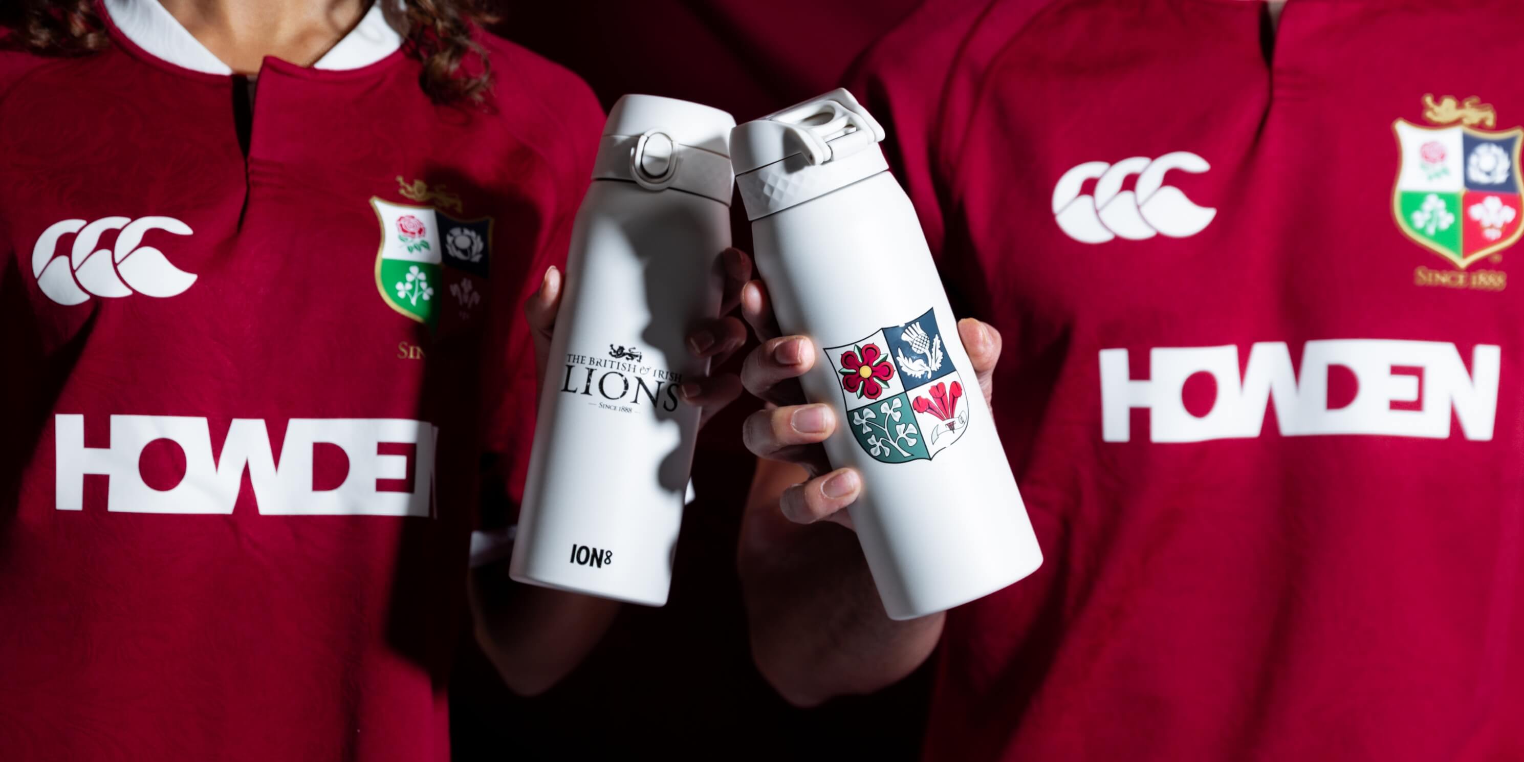 Two individuals, wearing red rugby jerseys with "HOWDEN" and the British & Irish Lions logo, hold up white ION8 water bottles featuring the Lions crest. The British & Irish Lions - Since 1888 is printed on one bottle.