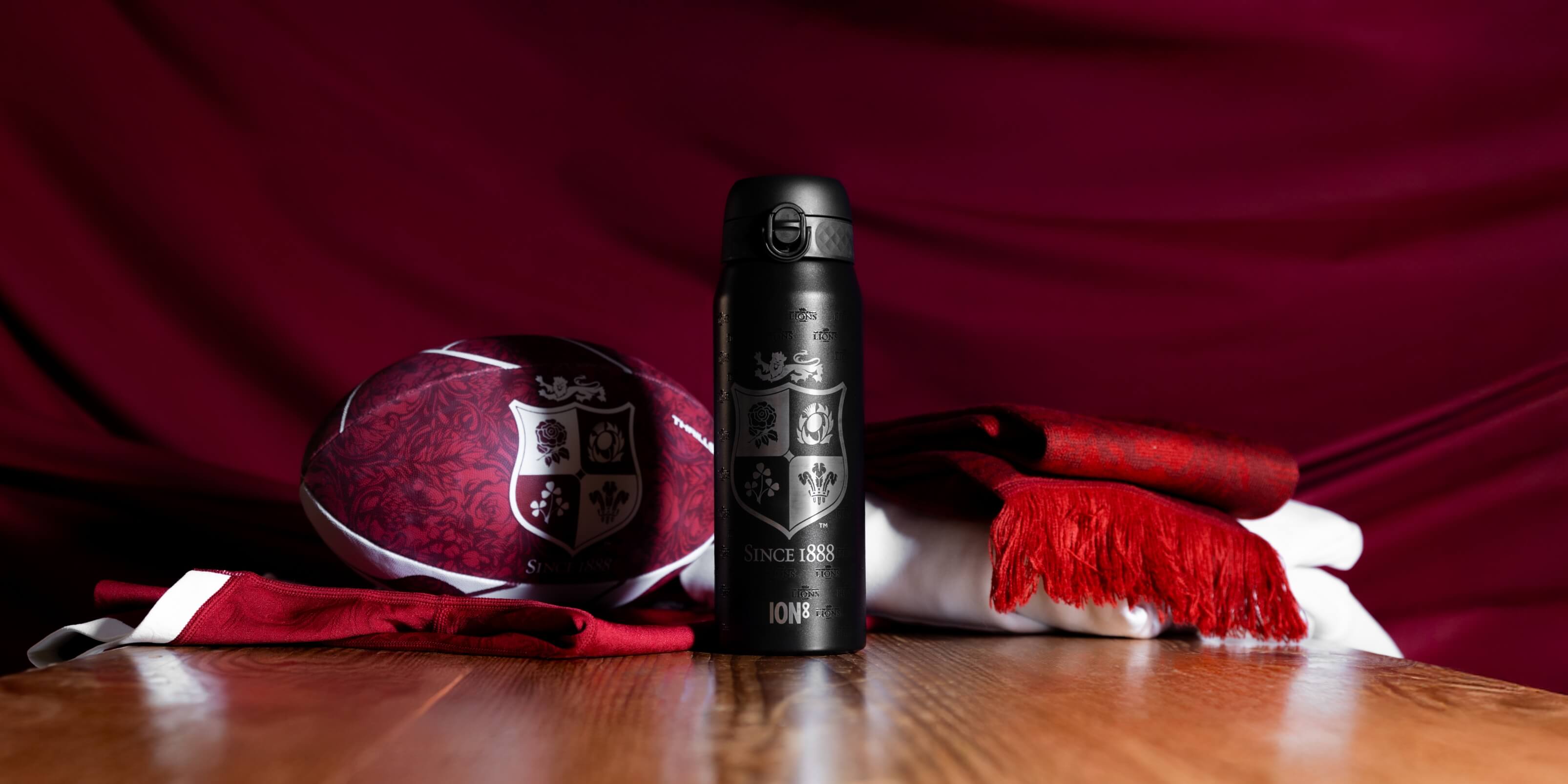 A black water bottle stands among a rugby ball, jersey, and scarf on a wooden surface; a dark red backdrop provides contrast. The bottle is engraved with "SINCE 1888" and "ION8".