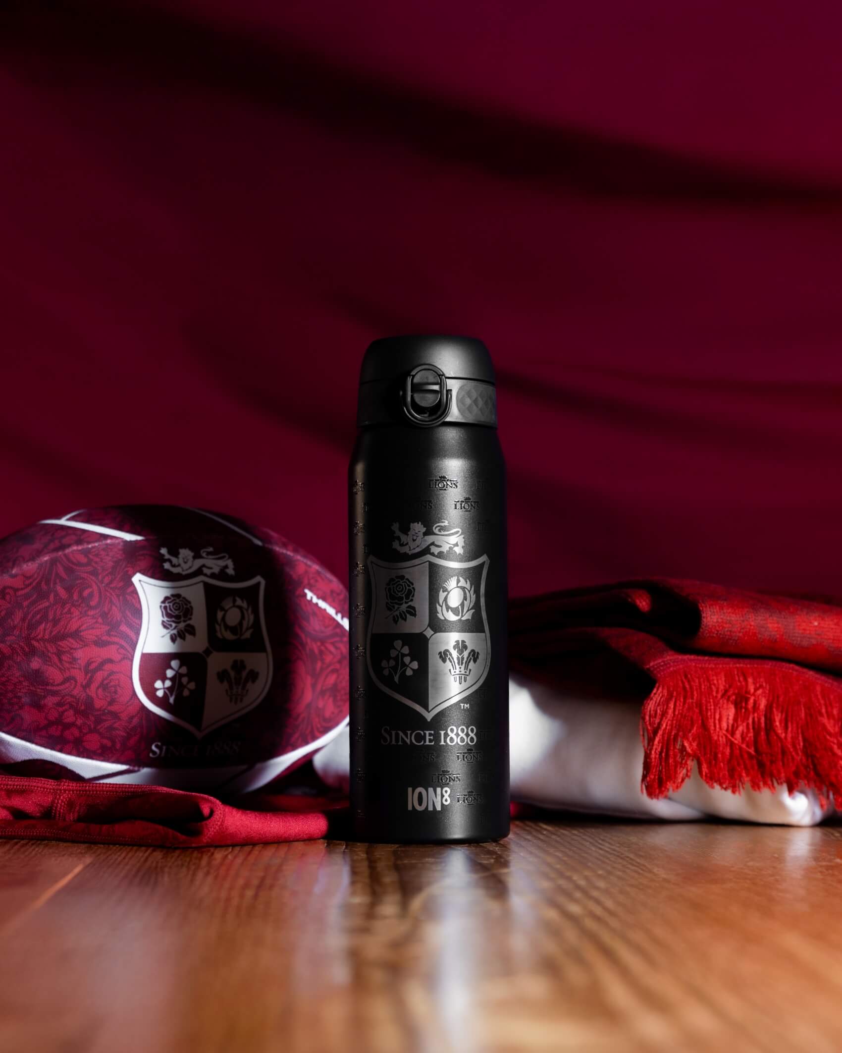 A black water bottle, featuring a crest and "SINCE 1888" and "ION8", stands on a wooden surface amidst a rugby ball and red scarf.